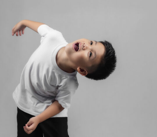 A child in a white t-shirt and black pants leans back with an open mouth, looking upwards against a plain grey background.