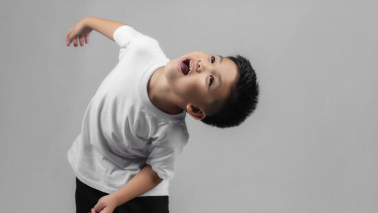 A child in a white t-shirt and black pants leans back with an open mouth, looking upwards against a plain grey background.